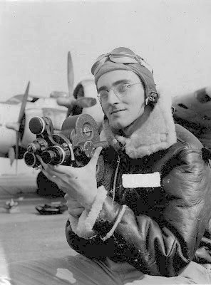  Close up view of Al Saldarini in wire-rimmed glasses wearing USAAF insulated flight jacker, and gunner's goggles atop a soft leather helmet. He's holding a 3-lensed camera.  Wing and two engine cowls and propellers are visible above his shoulders.